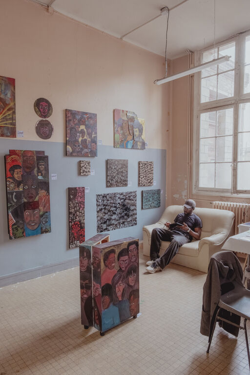 Salle 1 du Labo Victor Hugo. Exposition au Labo Victor Hugo à Rouen. Plusieurs peintures figuratives sont accrochées aux murs tandis qu’une personne est assise dans un fauteuil, travaillant sur une tablette à la lumière d’une grande fenêtre.