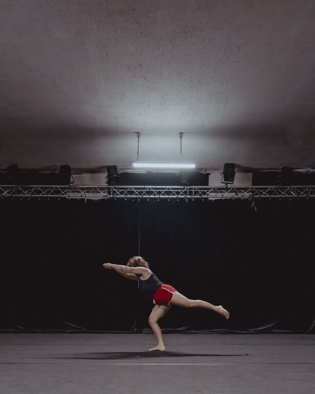 Salle 3 du Labo Victor Hugo. Danseuse exécutant une figure d’équilibre sur une scène de répétition du Labo Victor Hugo, devant un fond noir et sous un éclairage de travail.