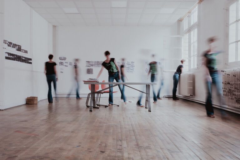 Salle 9 du Labo Victor Hugo. Vue d’un atelier collectif au Labo Victor Hugo. Plusieurs silhouettes floues se déplacent autour d’une table centrale, évoquant une phase de montage, de création ou de médiation artistique.