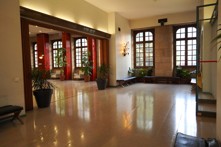 Fumoir du premier étage de la Halle aux Toiles avec larges fenêtres, bancs et plantes d’intérieur disposés dans un espace d’accueil calme et dégagé.
