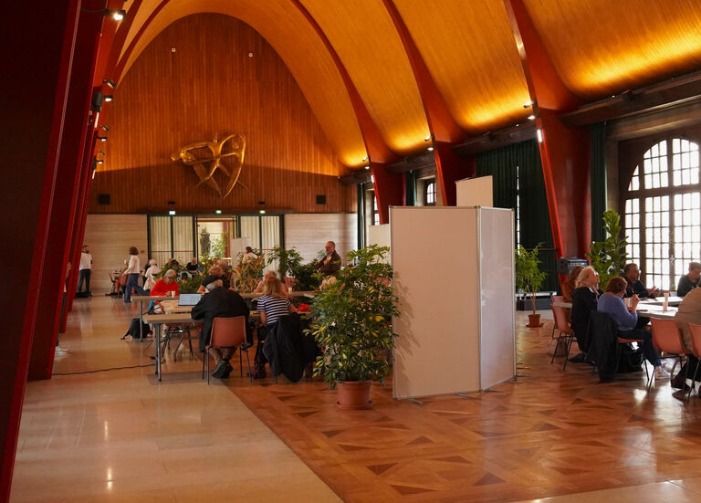 Salle polyvalente du premier étage de la Halle aux Toiles, où plusieurs groupes de personnes sont réunis autour de tables sous une haute voûte en bois.