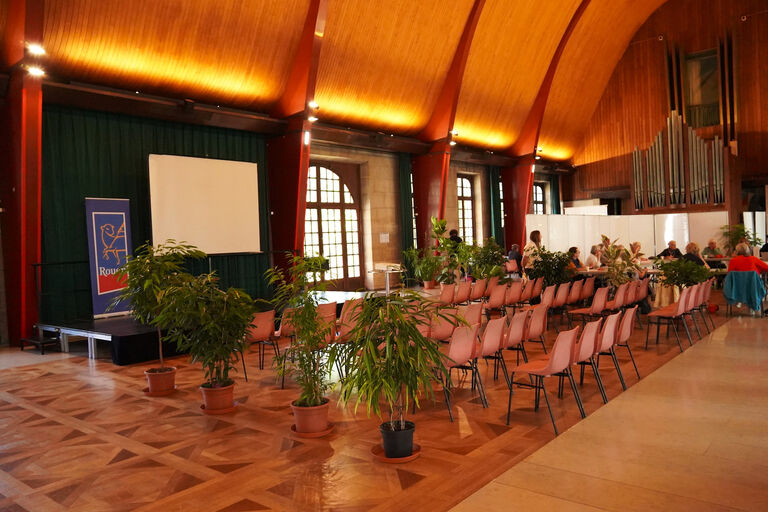 Salle polyvalente du premier étage de la Halle aux Toiles aménagée pour un événement, avec une scène équipée d’un écran de projection.