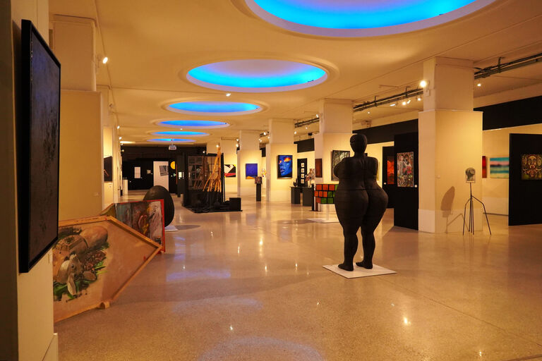 Hall d’exposition du rez-de-chaussée de la Halle aux Toiles présentant des œuvres d’art contemporaines et des sculptures, sous un plafond ponctué de puits de lumière circulaires bleutés.