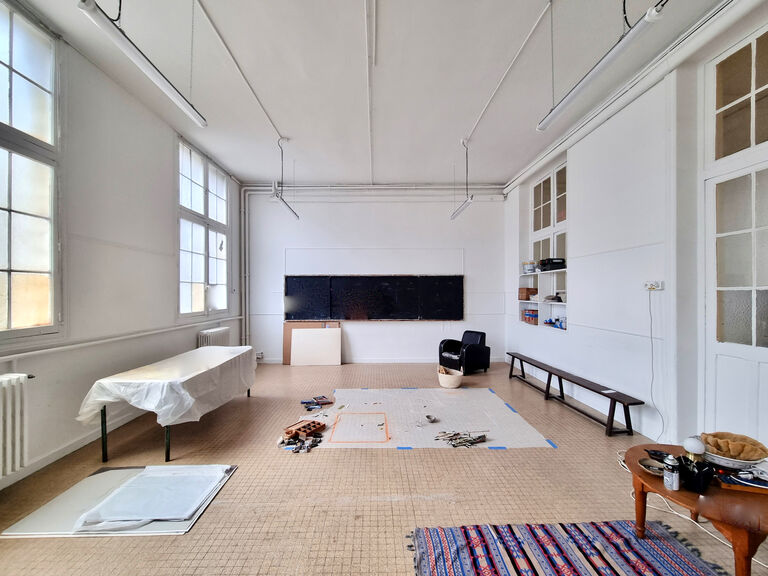 Salle lumineuse du Labo Victor Hugo aux murs blancs et aux hautes fenêtres, avec un espace de travail central protégé au sol et du matériel de création artistique.