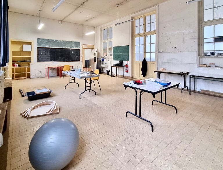 Grande salle d’atelier du Labo Victor Hugo avec hauts plafonds, grandes fenêtres, tableaux noirs muraux, tables de travail et matériel artistique disposés dans l’espace.