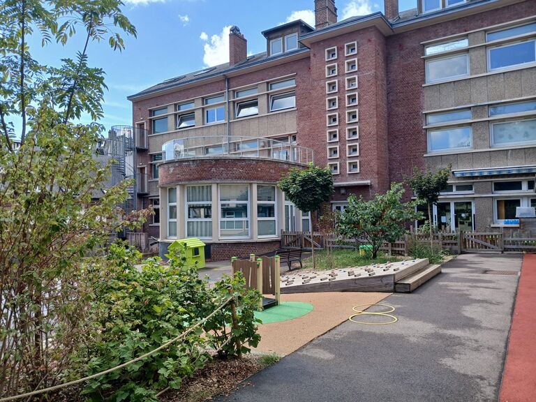 Vue extérieure d'une cour de récréation pavée et aménagée devant un grand bâtiment scolaire en briques rouges et béton. À gauche, une structure arrondie vitrée en demi-cercle soutient un balcon à l'étage. Au centre, la cour comprend une petite maisonnette en plastique vert, une structure d'escalade en bois sur un sol souple de sécurité et un plan incliné doté de prises d'escalade colorées. Des arbres, des buissons verdoyants et une barrière en bois bordent l'espace de jeux sous un ciel bleu.