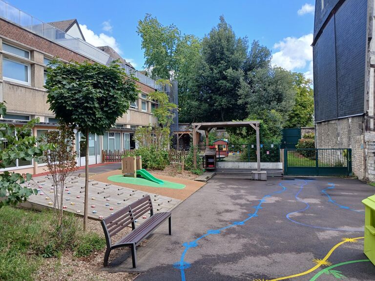 Vue d'un autre angle de la même cour de récréation, mettant en avant un banc en bois et métal au premier plan. Sur la gauche, le plan incliné d'escalade au sol fait face à une petite structure de jeu en bois équipée d'un toboggan vert. Au sol, le bitume est décoré de lignes peintes bleues et jaunes serpentant à travers la cour. À l'arrière-plan, une pergola en bois abrite du mobilier, jouxtant une barrière métallique verte et une végétation dense d'arbres hauts sous un ciel dégagé.