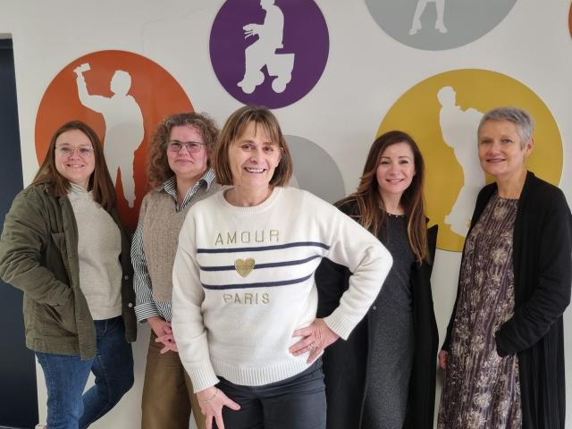 Cinq femmes de tout âge posent debout devant un mur blanc sur lequel il y a des ronds colorés en jaune, en orange et en violet 