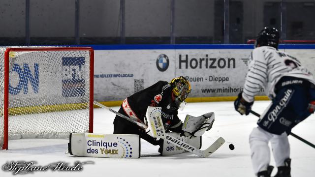 Le gardien de but de Rouen, en maillot noir, est à genoux devant sa cage et arrête le palet lancé par le joueur d'Angers en maillot blanc à rayures bleues  