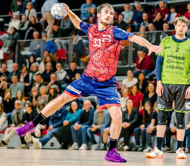 Le joueur de handball de Rouen en maillot rouge et en short bleu est en position de tir en extension vers la cage adverse