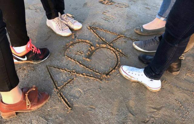 des personnes sont sur une plage et entourent trois lettres A, B et A tracées en majuscule sur le sable 