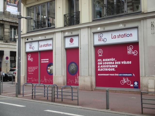 Vue de la rue Jeanne d'Arc de la vélo station Vue du trottoir d'en face de la façade de la vélo station rue Jeanne d'Arc avec une vitrine dotée d'affiches roses plastifiées sur lesquelles est écrit "ici, bientôt, on louera des vélos électriques"